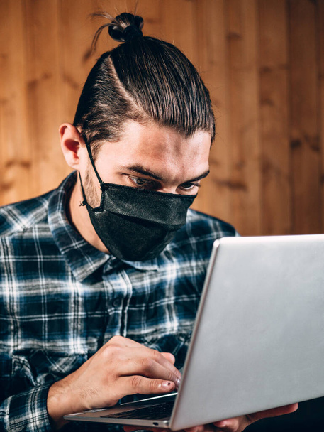 Um belo jovem freelancer com uma máscara protetora preta trabalha em um laptop prateado em casa. Operação remota durante a quarentena, para imprimir o texto e realizar negócios importantes. - Foto, Imagem