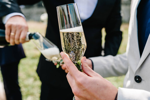 The groom pours champagne into a glass on the background of the friends on nature at the wedding ceremony. Close up. Toast. - Foto, Imagen