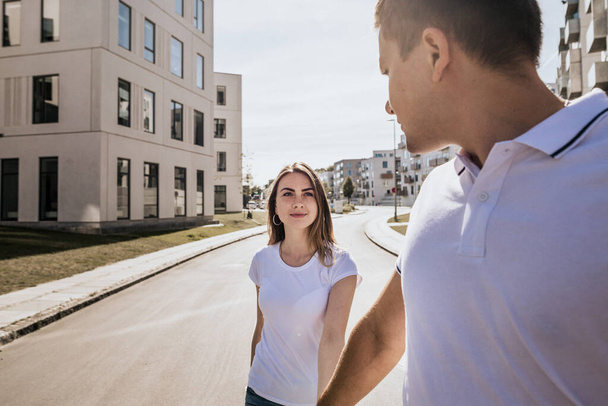 Close up of stylish couple in white T-shirt walking outdoors. Stylish loving family hold hands on building background - Photo, Image