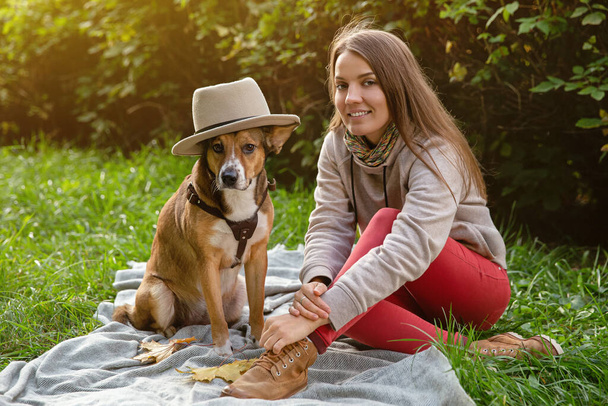 Autumn walk with your pet. A girl with dog in hat are sitting on plaid. - Photo, Image