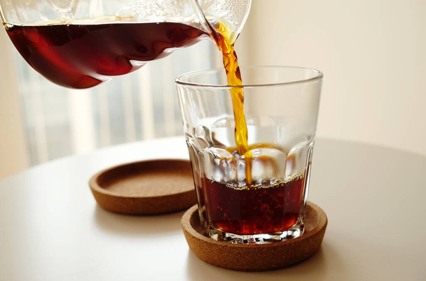 Μαύρο φίλτρο καφέ χύνει από μια κανάτα σε ένα ποτήρι από κοντά. Εξειδικευμένο τρίτο κύμα έννοια. Φωτεινό φόντο - Φωτογραφία, εικόνα