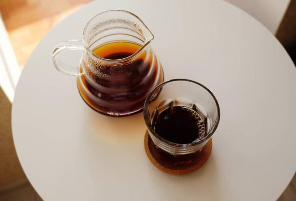 Μαύρο φίλτρο καφέ σερβίρεται σε κανάτα και ένα ποτήρι σε λευκό στρογγυλό τραπέζι. Στην κορυφή. Εξειδικευμένο τρίτο κύμα έννοια. Φωτεινό φόντο - Φωτογραφία, εικόνα