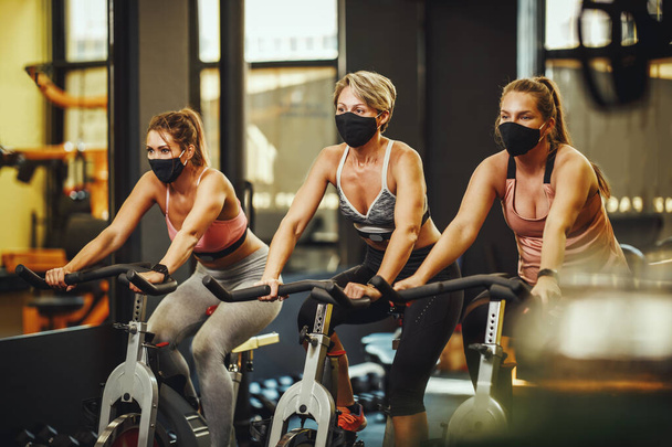 Grupa trzyosobowych przyjaciółek w masce ochronnej trenujących na rowerach treningowych w siłowni podczas pandemii Covid-19. - Zdjęcie, obraz