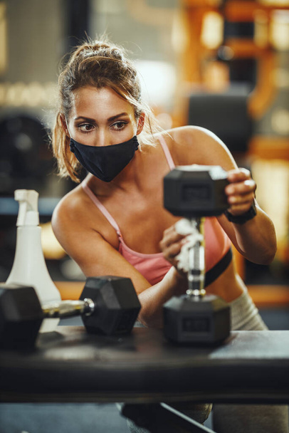 Prise de vue d'une jeune femme musclée avec masque protecteur nettoyant l "équipement de fitness après l'entraînement pendant la pandémie de Covid-19. - Photo, image