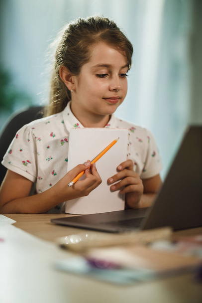 Egy szorgalmas kislányról készült felvétel, aki laptopot használ, hogy online leckét vegyen otthon a COVID-19 világjárvány idején.. - Fotó, kép