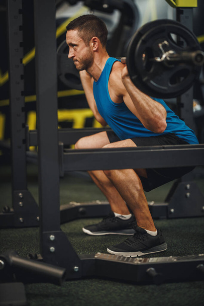 Aufnahme eines muskulösen Kerls in Sportbekleidung beim Training in der Crosslaufhalle. Er macht Kniebeugeübungen mit der Langhantel. - Foto, Bild