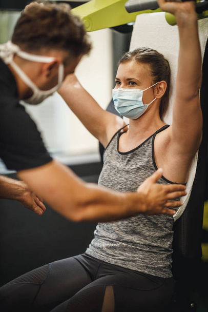 Shot of a muscular young woman with protective mask working out with personal trainer at the gym machine during Covid-19 pandemic. She is pumping up her shoulder muscule with heavy weight. - Photo, Image