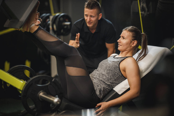 Aufnahme einer muskulösen jungen Frau in Sportbekleidung, die mit Personal Trainer im Fitnessstudio trainiert. Sie macht Übungen für ihre Beine an einem Beinpressgerät. - Foto, Bild