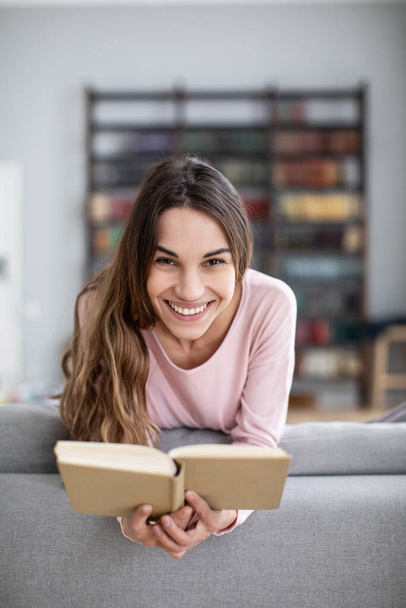 Attractive young woman is reading a book, looking at camera and smiling at home. - Foto, Imagem