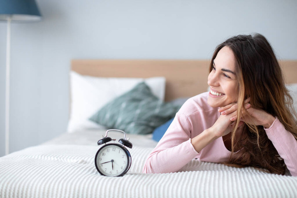 Smiling young woman lying awake in bed with alarm clock, good morning. Concept time management. - Φωτογραφία, εικόνα