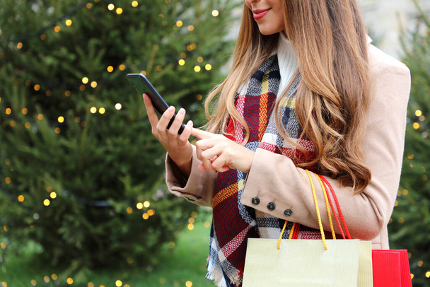 Όμορφη νεαρή ελκυστική γυναίκα που χρησιμοποιεί το smartphone για online αγορές και μεταφέρουν τσάντες για τα Χριστούγεννα - Φωτογραφία, εικόνα