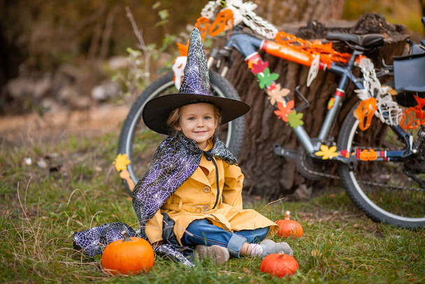 Маленька дівчинка на свято Хеллоуїна в природі, дитина біля велосипеда прикрашена гірляндами
 - Фото, зображення
