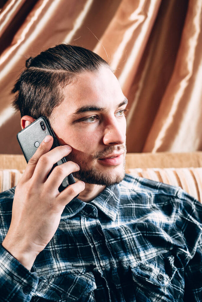 Un joven guapo con una camisa azul se comunica con un teléfono móvil y trabaja desde la comodidad de su casa durante la cuarentena. Trabajo remoto en línea. - Foto, imagen