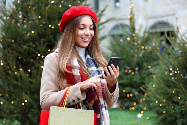 Portrait of smiling young shopper woman buying online on smartphone in city street with christmas trees on the background - Photo, Image