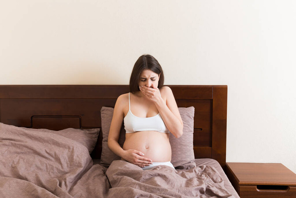 Schwangere leiden unter Übelkeit. Schwangerschaftssymptome, Erwartungstoxikose. Junge erbrechende Frau sitzt auf dem Bett. Konzept der Elternschaft. - Foto, Bild