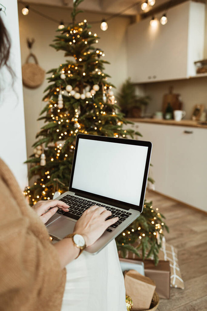 Młoda kobieta pracuje na laptopie z pustym ekranem wyświetlacza z miejsca kopiowania makiety. Nowoczesny salon wnętrz urządzone na Boże Narodzenie, Nowy Rok uroczystości z choinką. - Zdjęcie, obraz