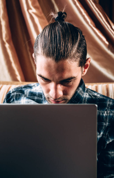 Un apuesto joven freelancer con una camisa azul trabaja en una laptop plateada en casa. Trabajo remoto durante la cuarentena. - Foto, imagen