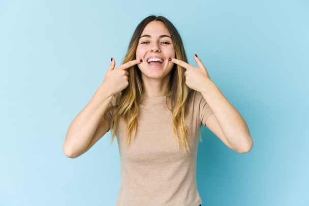 Young caucasian woman isolated on blue background smiles, pointing fingers at mouth. - Foto, afbeelding