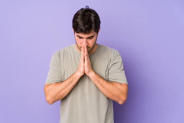 Young man isolated on purple background praying, showing devotion, religious person looking for divine inspiration. - Foto, Bild