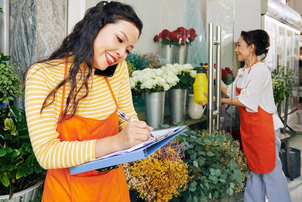 Lächelnde hübsche junge Floristin telefoniert mit Kunden und macht sich Notizen im Dokument - Foto, Bild