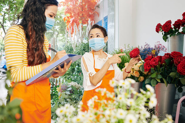 Team of florists in medical masks discussing which flowers to order for the next week and filling document - Photo, Image