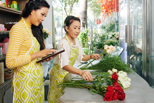 Proprietario del negozio di fiori che lavora sul computer tablet quando fiorista facendo bouquet per il cliente - Foto, immagini