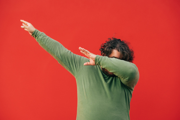 Σγουρομάλλης υπέρβαρος νεαρός άνδρας που δείχνει επιδεικτική χειρονομία σε κόκκινο φόντο. Χοντρός τύπος με πράσινη μπλούζα χορεύει νταμπ σε φόντο κόκκινου τοίχου.. - Φωτογραφία, εικόνα