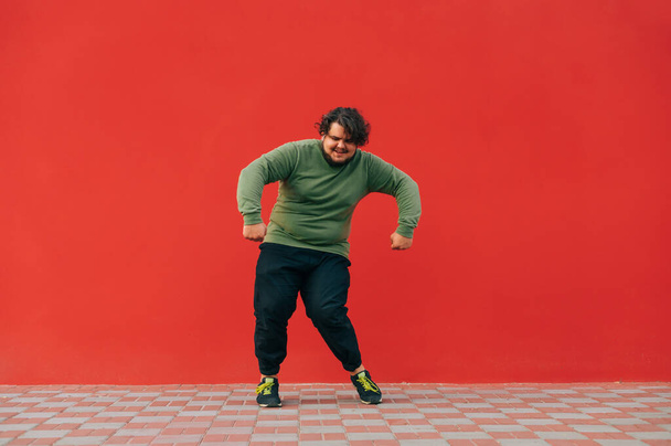 Charismatic positive fat man in casual clothes dancing on a red wall background. An overweight guy shows a dance performance on the street. - Photo, image