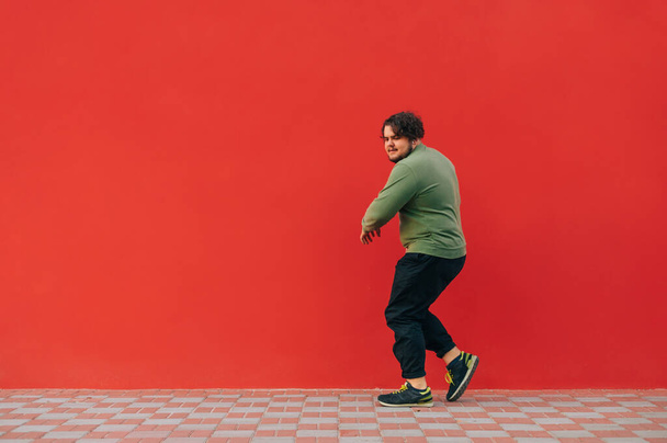 Grappige dikke man in groene sweater danst hip hop dansen op rode muur achtergrond en luistert naar muziek op koptelefoon. - Foto, afbeelding
