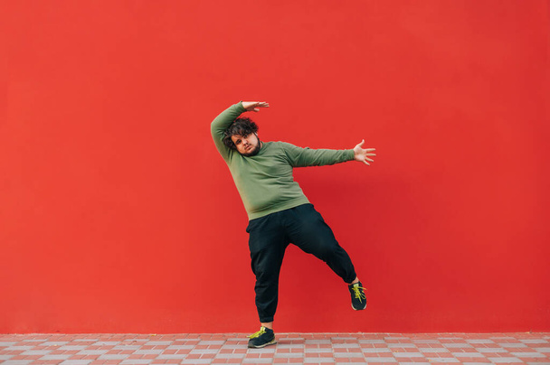 Grappige jongeman met overgewicht en krullend haar danst tegen de achtergrond van een rode muur, kijkt naar de camera en beweegt. - Foto, afbeelding