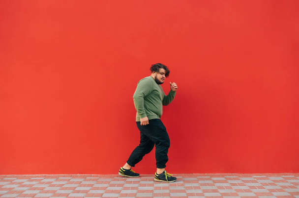 Elegante joven con sobrepeso en ropa casual escucha música en auriculares inalámbricos y baila sobre un fondo de pared roja, vista lateral. - Foto, imagen