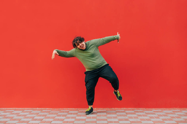 Šťastný mladý muž s nadváhou v neformálním oblečení tančí na červeném pozadí a dívá se na kameru s úsměvem tváře. Izolované. - Fotografie, Obrázek