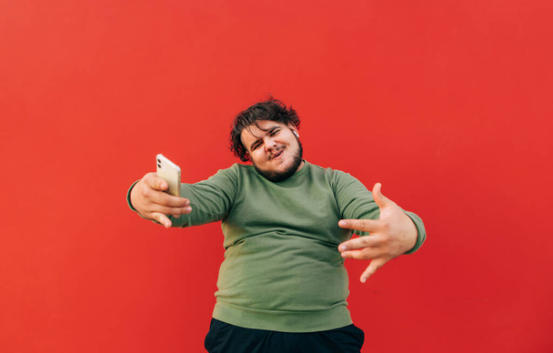 Portrét pozitivního kudrnatého muže s nadváhou na červeném pozadí, tančícího s úsměvem na tváři a dívajícího se do kamery. Tlusťoch poslouchá hudbu a tance, izolovaný. - Fotografie, Obrázek