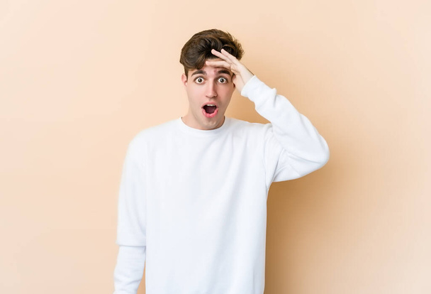 Young caucasian man isolated on beige background looking far away keeping hand on forehead. - Foto, Imagen