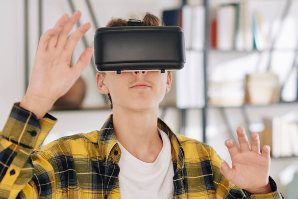 Tiener in geruite shirt spelen spel in virtual reality headset thuis - Foto, afbeelding