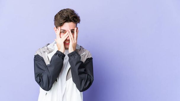 Young caucasian man isolated on purple background blink at the camera through fingers, embarrassed covering face. - Photo, Image