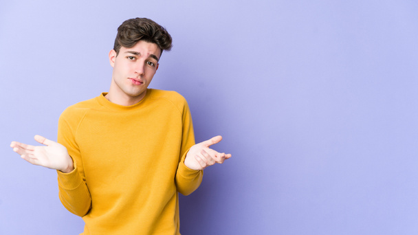 Young caucasian man isolated on purple background doubting and shrugging shoulders in questioning gesture. - Photo, Image