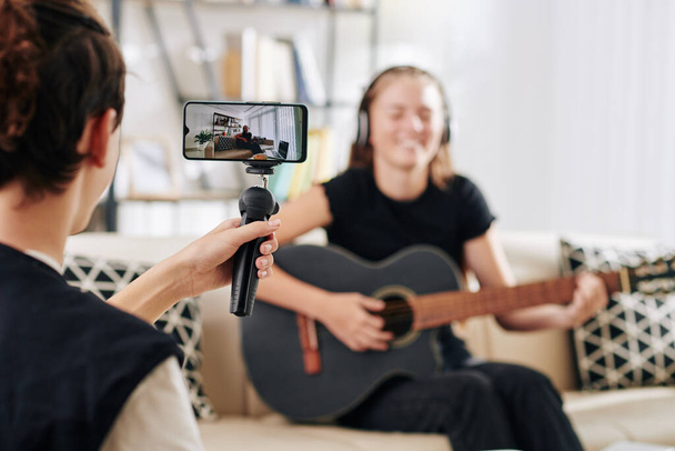 adolescent garçon à l'aide smartphone sur monopod lors du tournage de sa sœur ou ami jouer de la guitare et chanter chanson elle a écrit - Photo, image