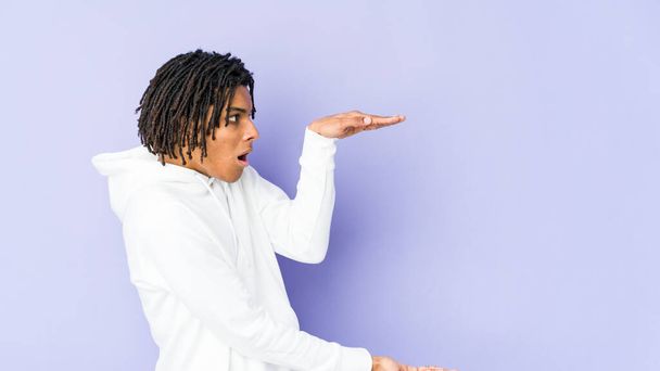 Νεαρός Αφροαμερικάνος ραστάς σοκαρισμένος και έκπληκτος κρατώντας ένα αντίγραφο χώρο μεταξύ των χεριών. - Φωτογραφία, εικόνα