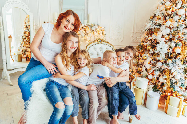 Ευτυχισμένη οικογένεια μητέρα και πέντε παιδιά χαλαρώνουν παίζοντας κοντά στο χριστουγεννιάτικο δέντρο την παραμονή των Χριστουγέννων στο σπίτι. Mom κόρες γιους στο φως δωμάτιο με διακόσμηση το χειμώνα. Χριστούγεννα Πρωτοχρονιά για γιορτή - Φωτογραφία, εικόνα