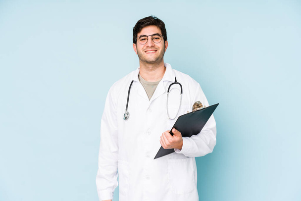 Mladý lékař muž izolovaný na modrém pozadí šťastný, usmívající se a veselý. - Fotografie, Obrázek