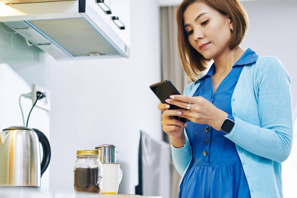 Красивая молодая азиатка, ждущая кофе на кухонном столе и отвечающая на смс-ки на смартфоне - Фото, изображение