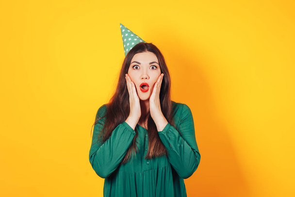 Woman with a surprised face holding her face with her hands in a green dress on a yellow background - Foto, Bild