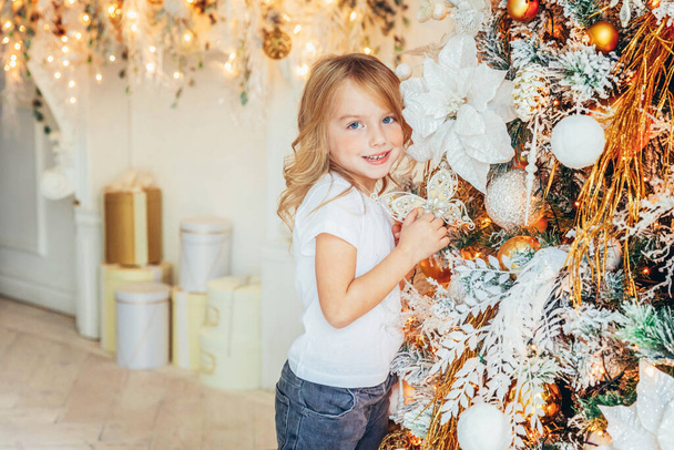 Little girl decorating Christmas tree on Christmas eve at home. Young kid in light room with winter decoration. Happy family at home. Christmas New Year december time for celebration concept - 写真・画像