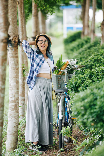 Szczęśliwa młoda Chinka stojąca w parku obok roweru z koszykiem zakupów w gaju lub parku - Zdjęcie, obraz
