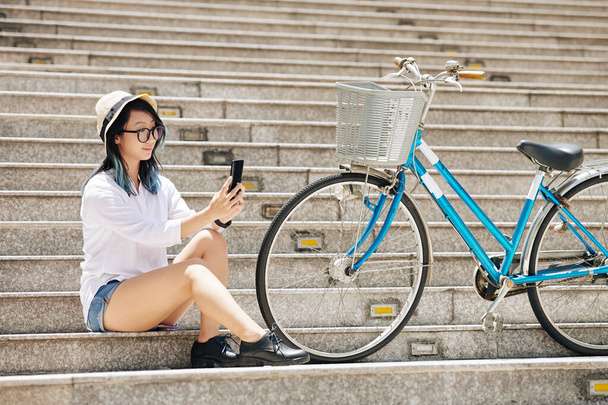 Bella giovane donna cinese seduta sui gradini accanto alla sua bicicletta e scattare selfie per i social media - Foto, immagini