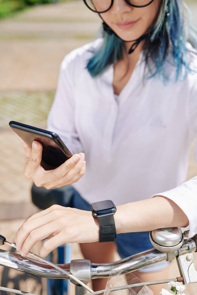 Νεαρή γυναίκα συγχρονισμό smartphone και smartwatch πριν από την οδήγηση ποδήλατο στην πόλη - Φωτογραφία, εικόνα
