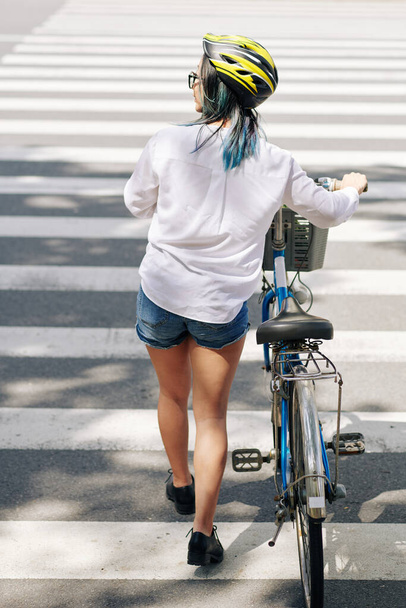 Νεαρή γυναίκα με λευκό πουκάμισο και denim σορτς που περπατά δίπλα στο ποδήλατο όταν διασχίζει το δρόμο - Φωτογραφία, εικόνα