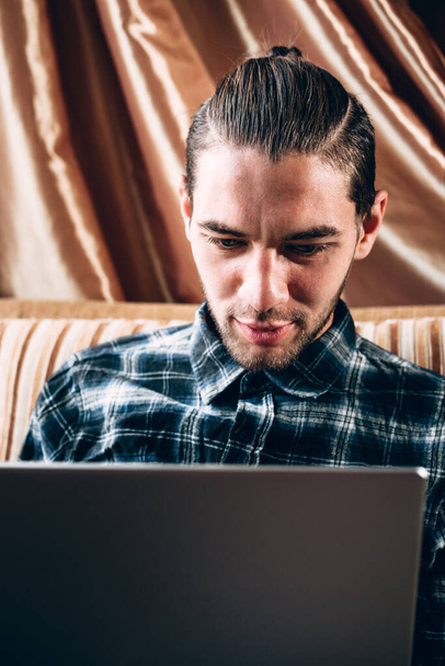Un apuesto joven freelancer con una camisa azul trabaja en una laptop plateada en casa. Trabajo remoto durante la cuarentena. - Foto, Imagen