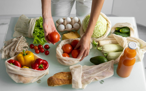 Womans hand, holding a reusable grocery bag with vegetables on a kitchen at home and takes tomato out. Zero waste and plastic free concept. Mesh cotton shopper with vegetables. Ecology. - Foto, immagini